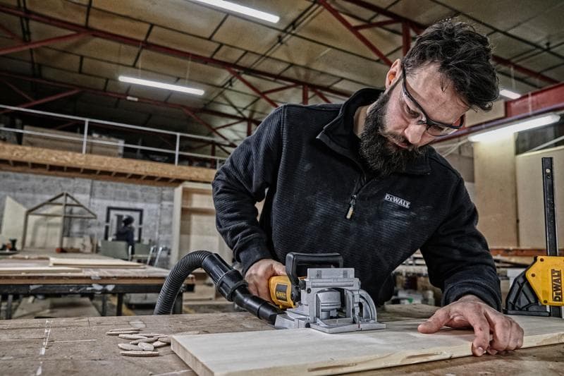 Una persona utiliza una herramienta de carpintería DEWALT DW682K para cortar una pieza de madera sobre una mesa en un taller. El rostro de la persona está oculto por privacidad.