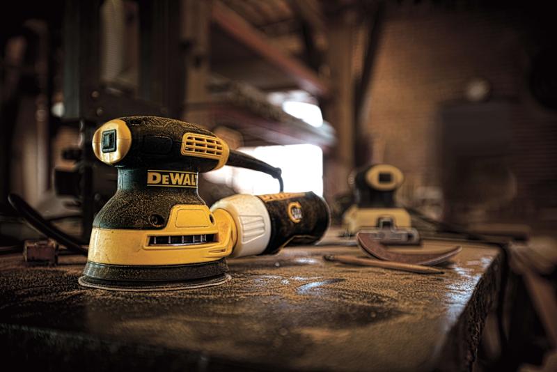 Primer plano de una lijadora eléctrica DEWALT DWE6421 sobre un banco de trabajo cubierto de polvo en un taller, con discos de lijado y otra lijadora visibles al fondo.
